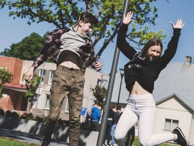 Two teenagers joyfully jumping outdoors in Šiluva, Lithuania under a bright blue sky. Captured in springtime.